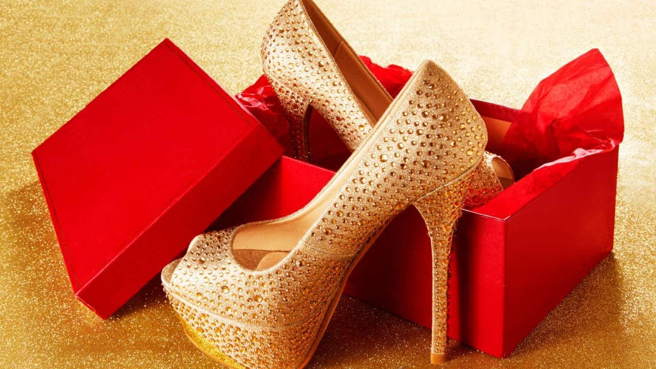 Um par de sapatos dourados de salto alto saindo de uma caixa de presente vermelha.