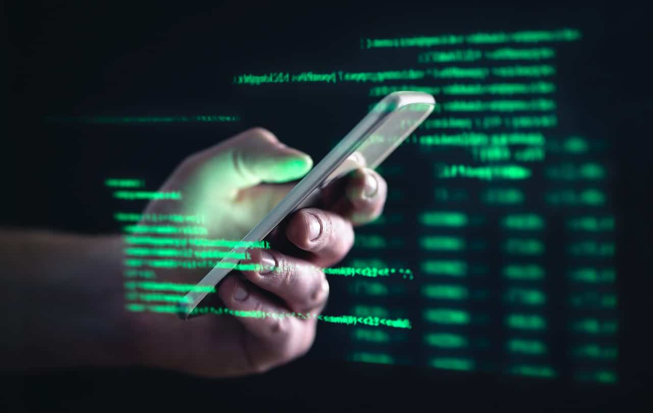 Imagem de uma mão de pessoa branca pegando um celular cinza claro, simbolizando um celular clonado. O fundo é preto com letras pequenas verdes