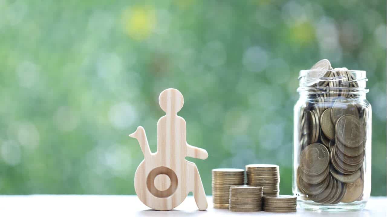 Imagem de cadeirante ao lado de uma pilha de moedas representando o auxílio-inclusão.