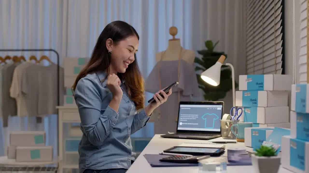 Mulher sorrindo para celular em um escritório com materiais de trabalho, como notebook, tablet, luminária tudo isso sobre uma mesa branca