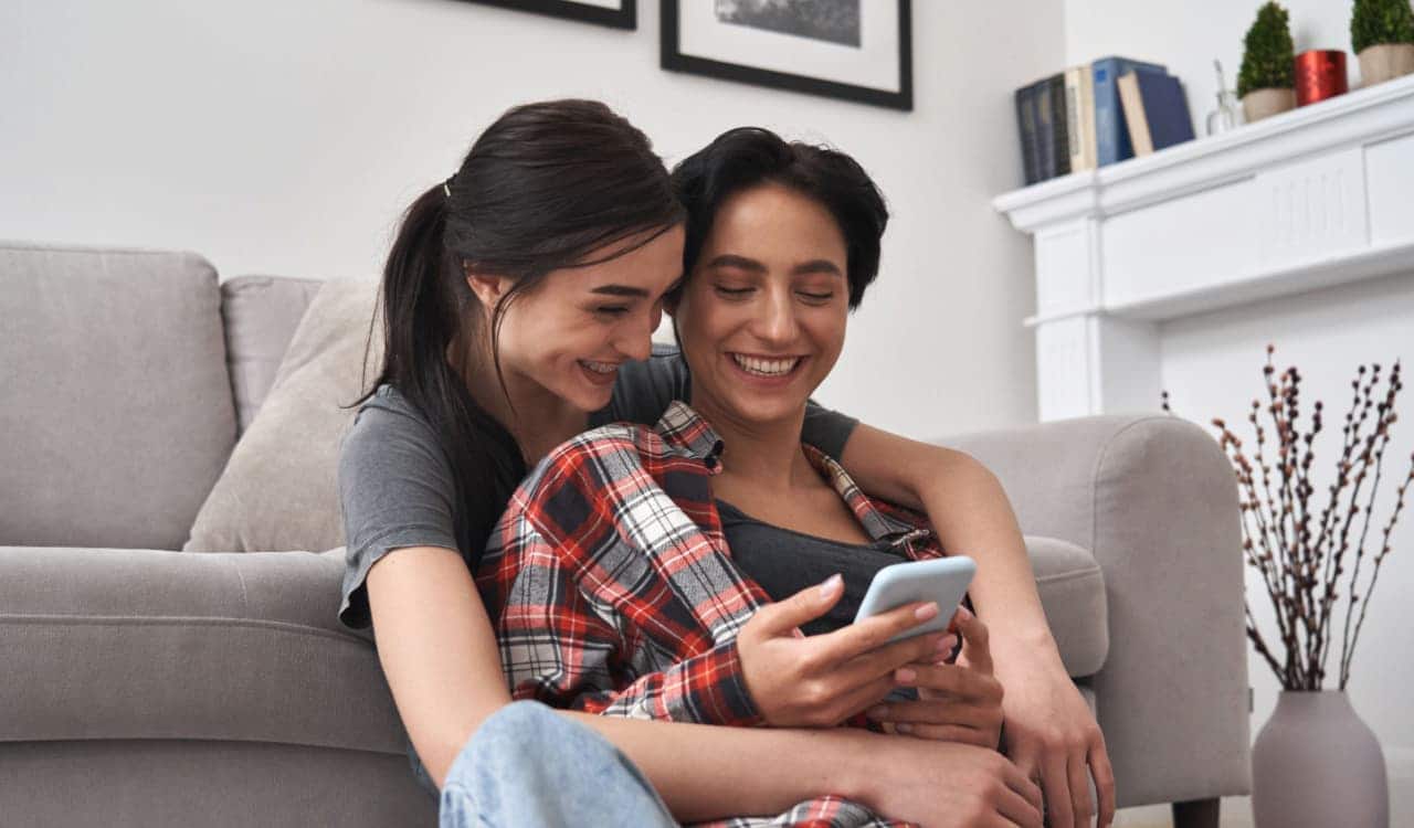 Duas mulheres abraçadas olhando para tela do celular e sorrindo