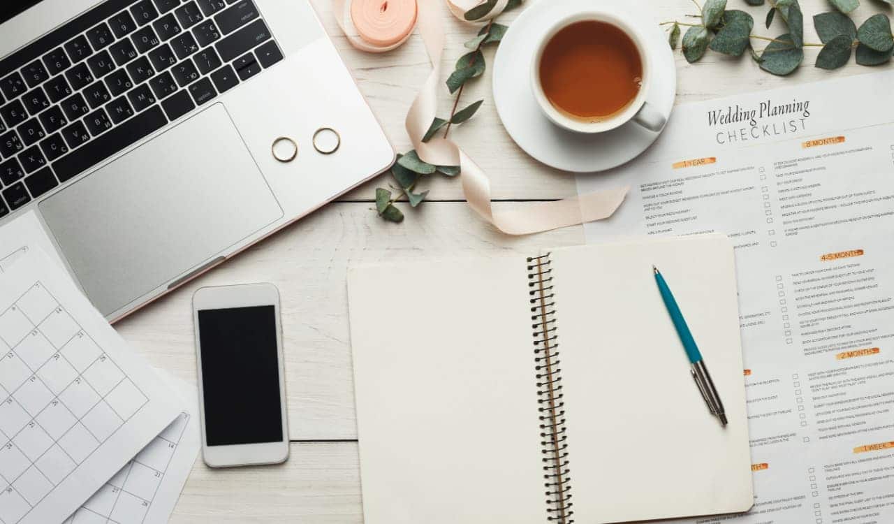 Caderno de planejamento, celular, notebook, xícara de chá e alianças de casamento espalhados sobre uma mesa