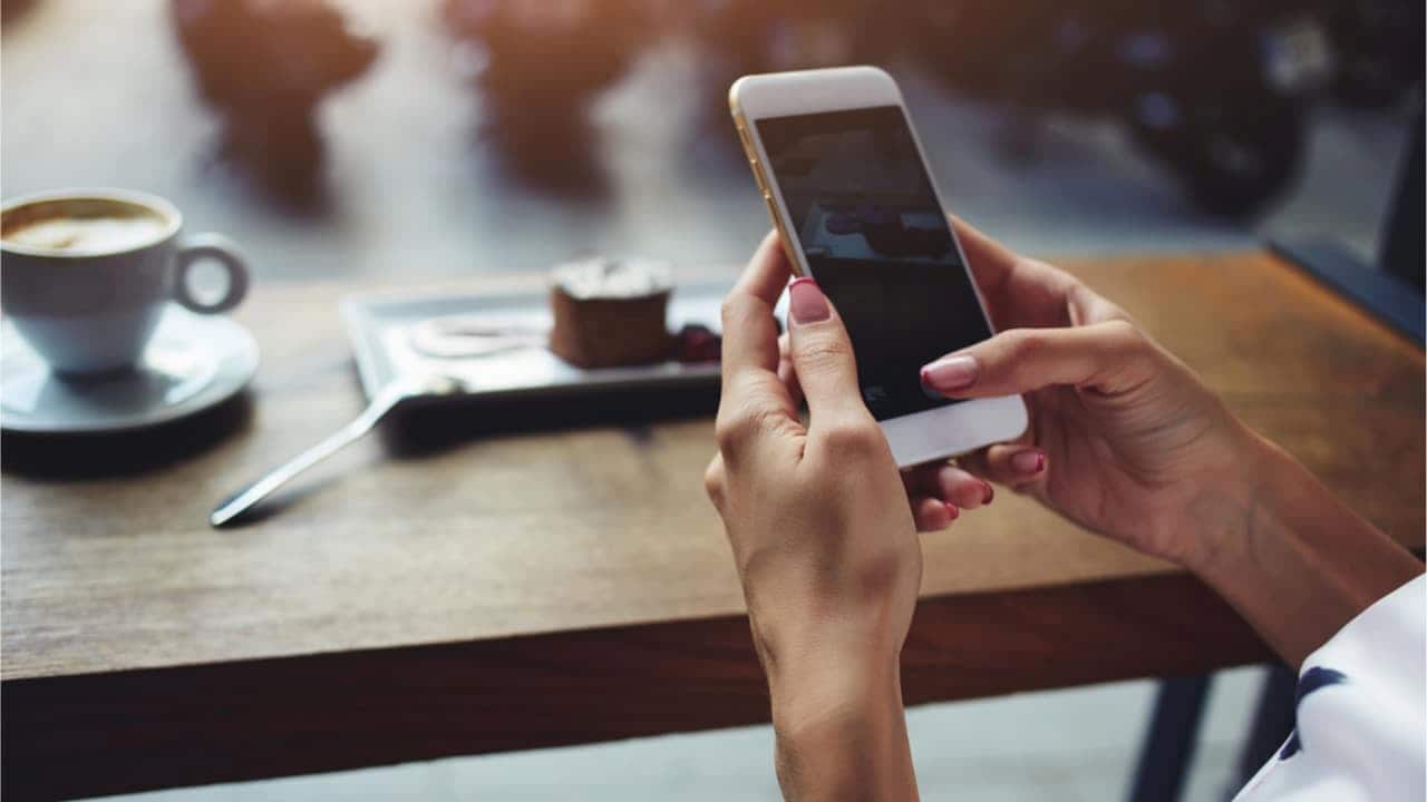Mãos segurando celular e mesa com xícara de café desfocada ao fundo empréstimo propaganda celular seguro