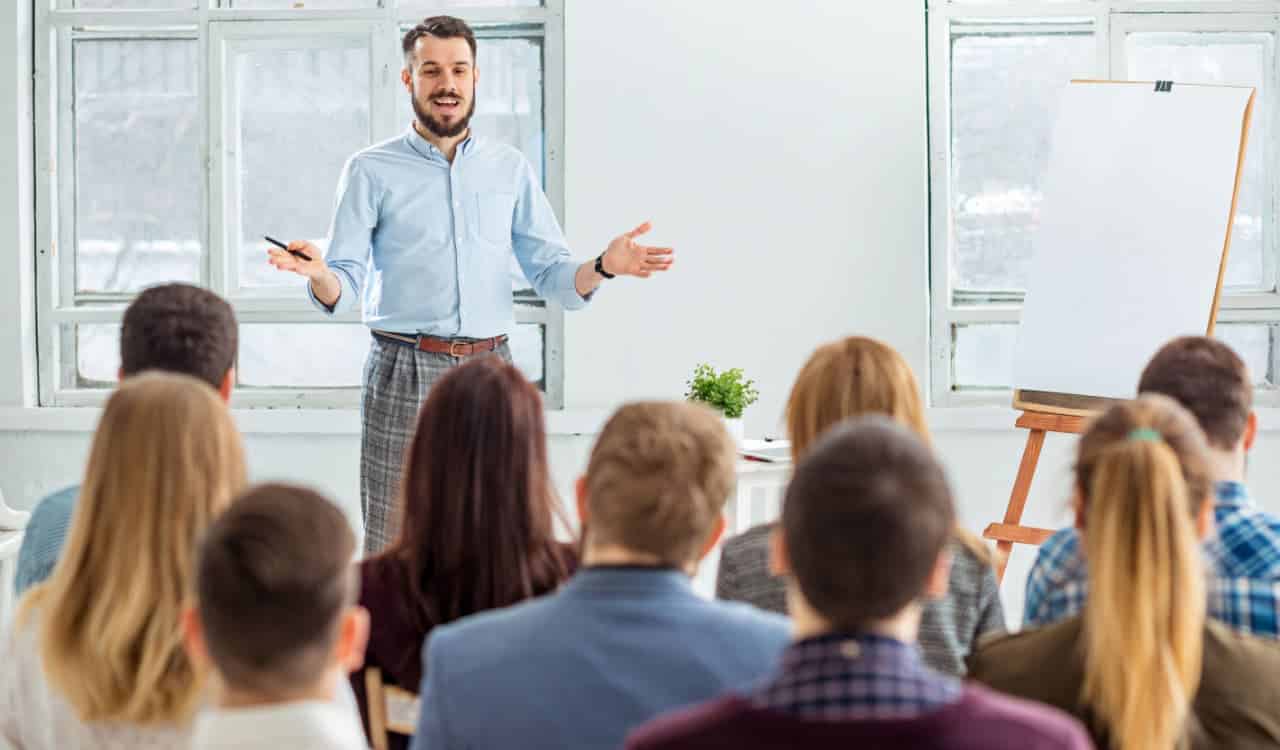 Homem em pé falando com diversas pessoas que estão sentadas à sua frente