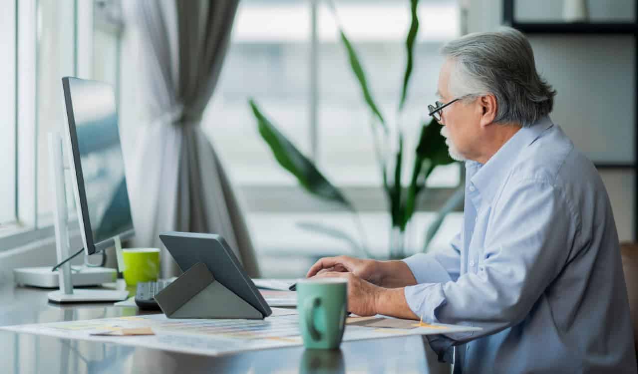 Homem idoso trabalhando em computador. Sobre a mesa, várias folhas espalhadas.