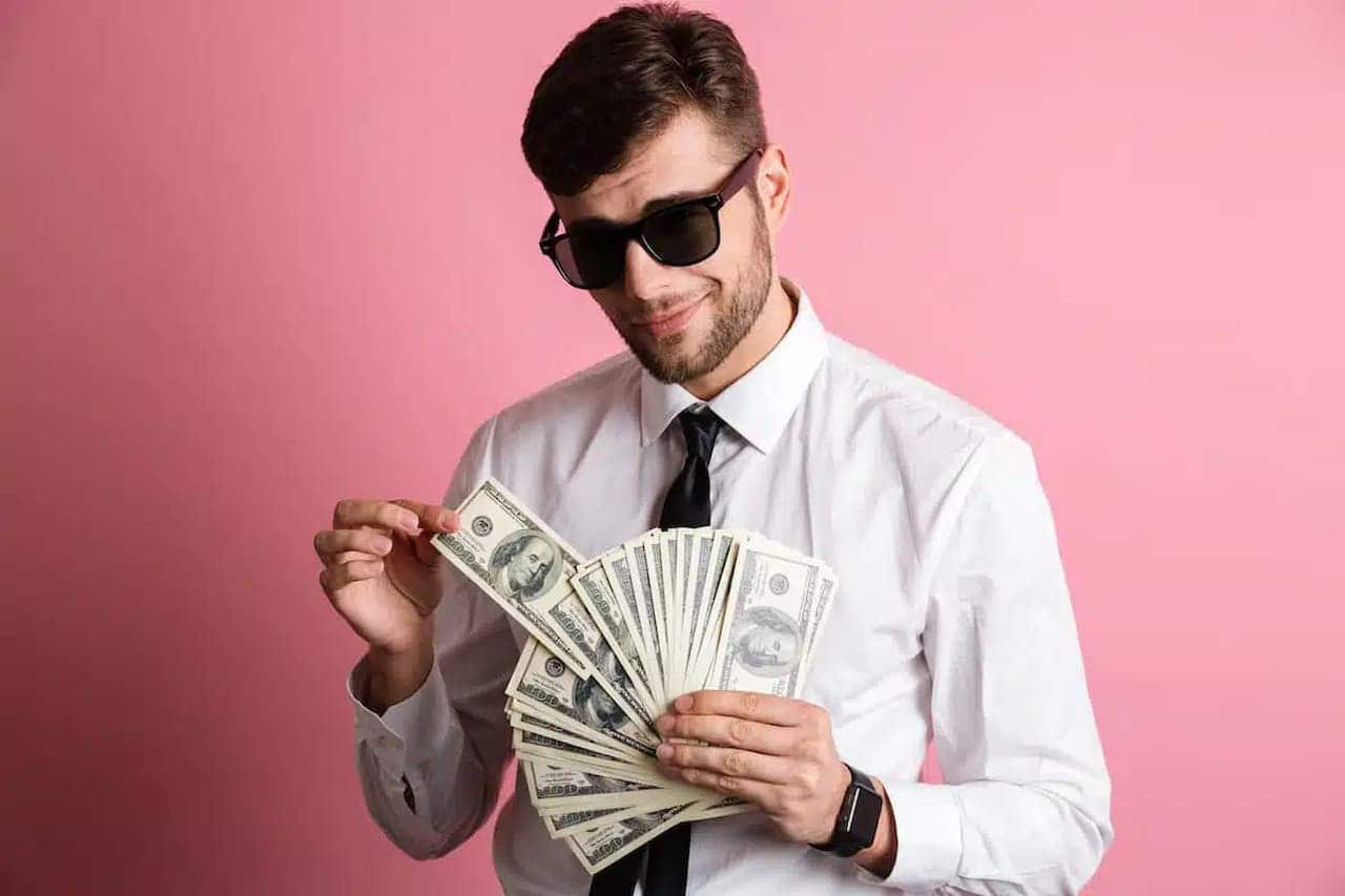 A imagem mostra um homem segurando vários dólares, com óculos escuro e sorridente.