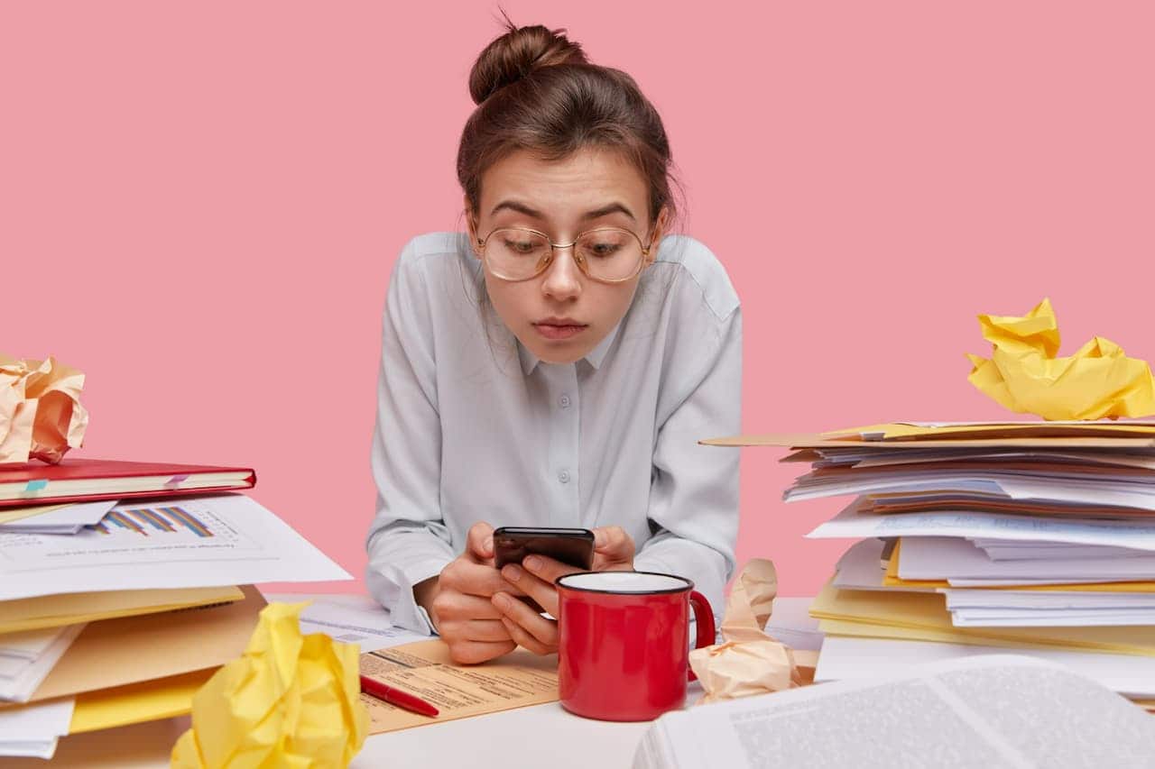 Mulher mexendo no celular sentada na frente de mesa cheia de pastas e papéis do trabalho procrastinação