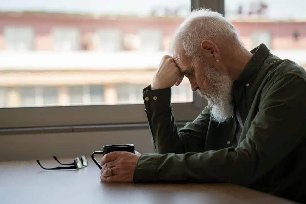 A imagem mostra um homem idosos, com a cabeça baixa, apoiada em uma das mãos, aparentemente triste
