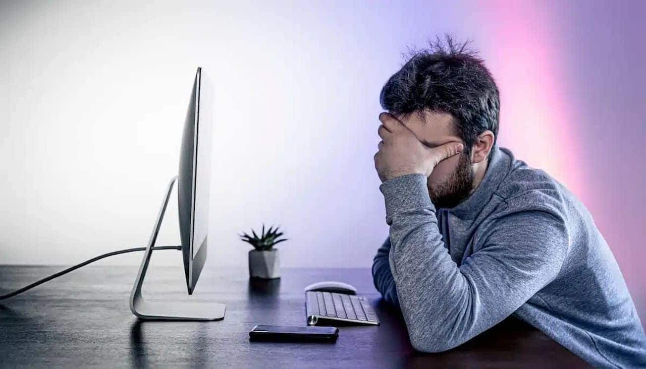 Homem aparentemente exausto. Com uma das mãos cobrindo a região dos olhos, em frente ao computador
