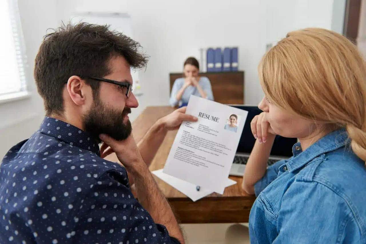 Dois recrutadores analisando o currículo de um candidato, que espera ao fundo, durante uma entrevista de emprego.