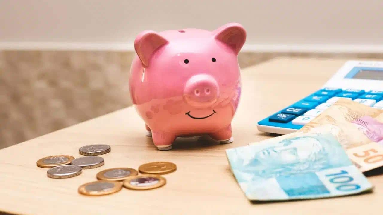 Cofrinho em formato de porco ao lado de várias moedas, uma nota de 100 reais e uma calculadora.