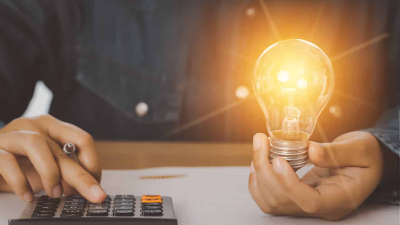Homem digitando em calculadora enquanto segura uma lâmpada acessa na outra mão