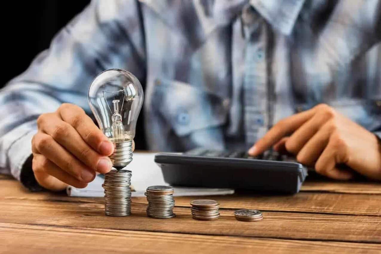 Homem com uma lâmpada em cima de uma pilha de moedas, enquanto usa a calculadora
