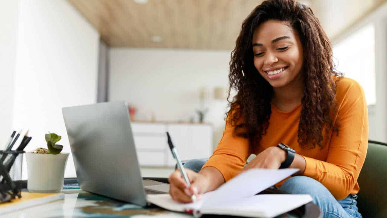 Mulher estudando em uma mesa, usando um notebook enquanto faz anotações no caderno e sorri.
