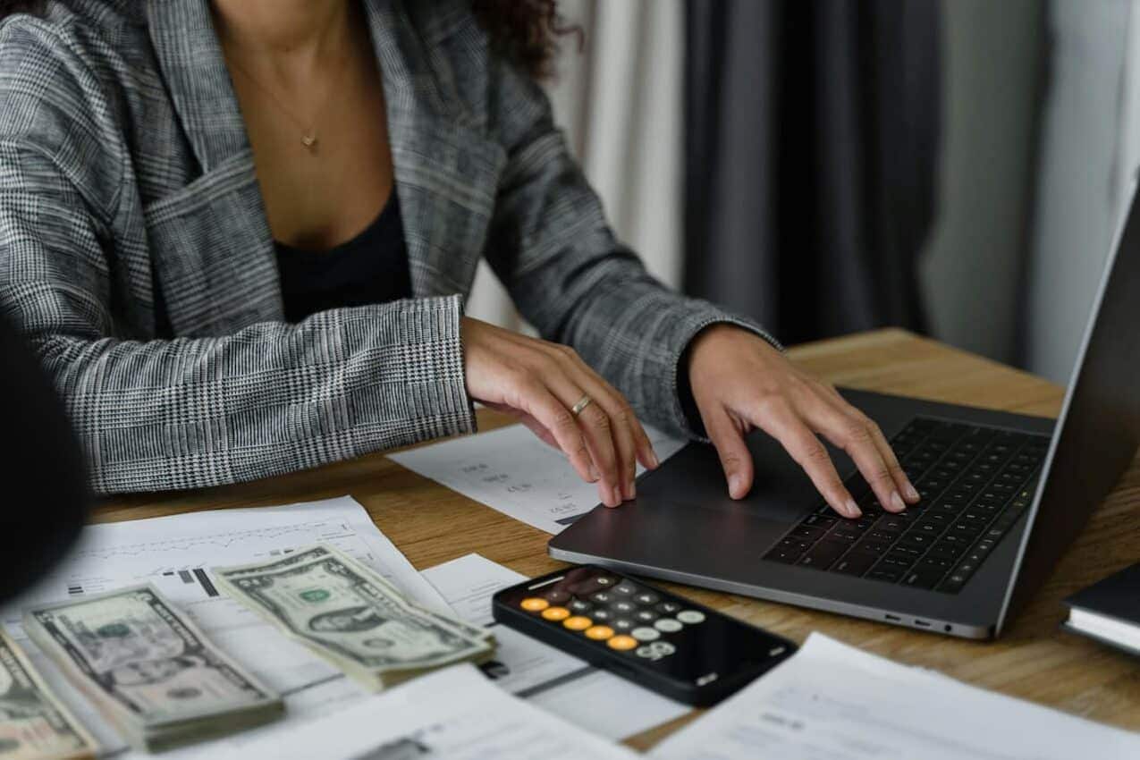 Imagem de uma mulher mexendo em um notbook, ao lado tem um celular com a calculadora aberta na tela e dinheiro de papel