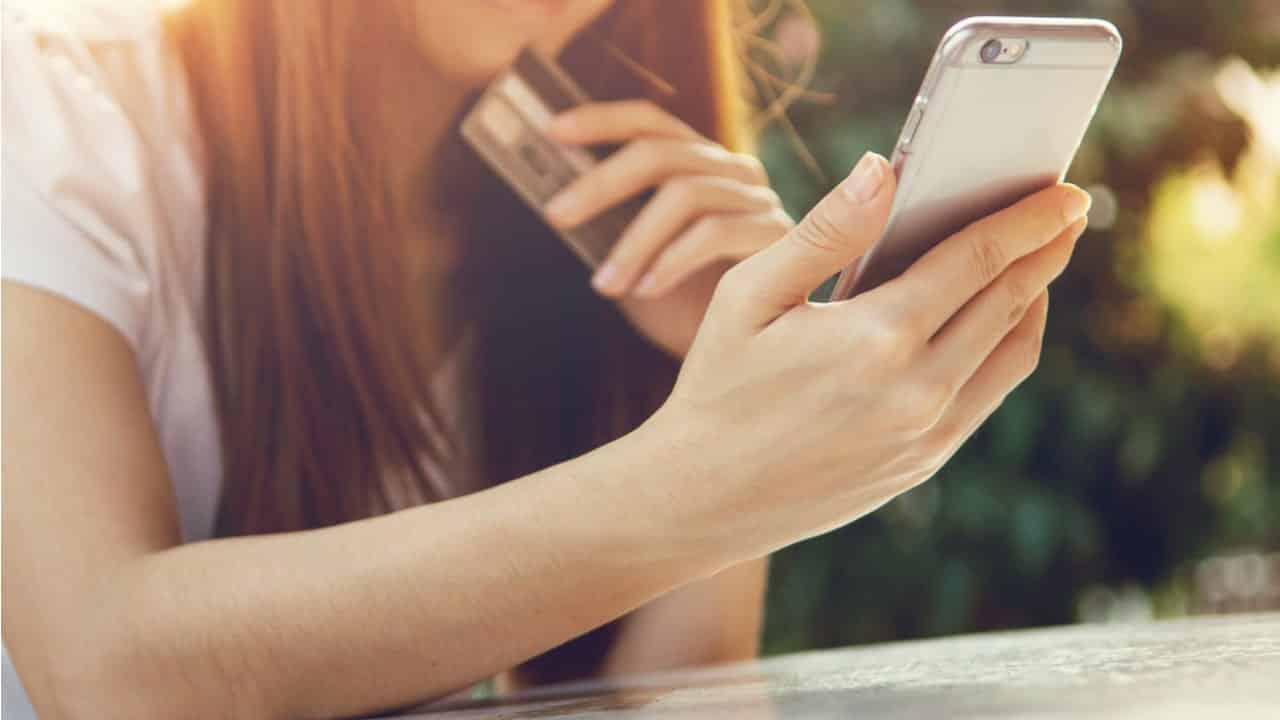Mulher segurando e olhando para o celular, enquanto a outra mão segura o cartão de banco.