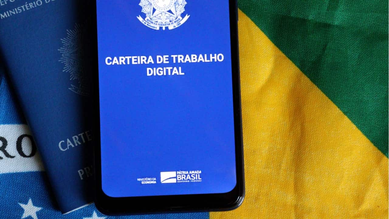 Celular com app da Carteira de Trabalho Digital sobre uma Carteira de Trabalho física e uma bandeira do Brasil.