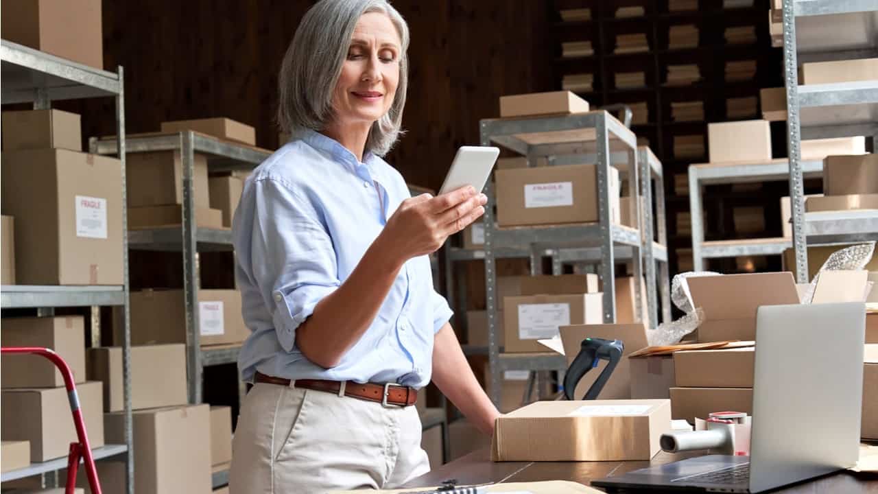 Senhora com celular na mão em um depósito de mercadoria trabalhando.