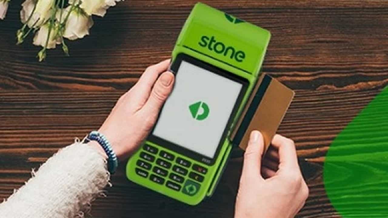 Mãos inserindo cartão na maquininha verde da Stone, sobre superfície/fundo marrom