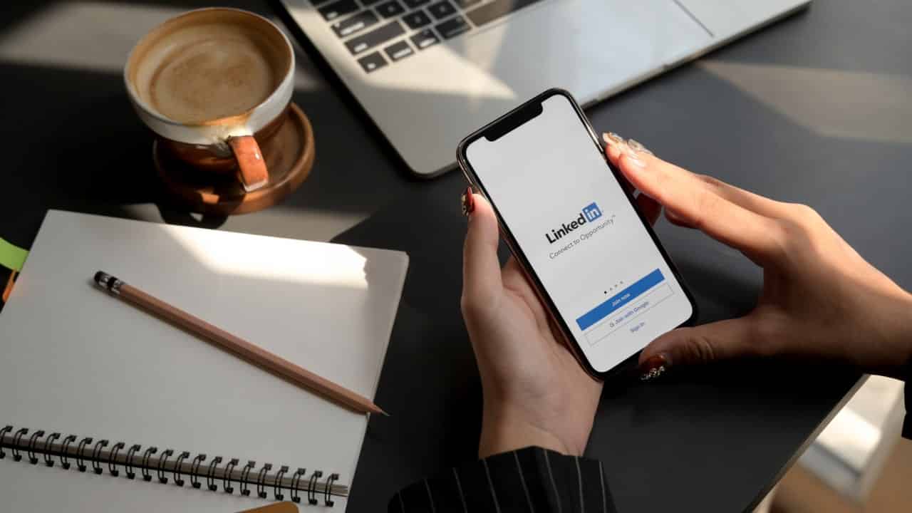 Pessoa acessa o LinkedIn pelo celular, ao redor há uma caneca com café, um notebook e um bloco de notas com um lápis em cima