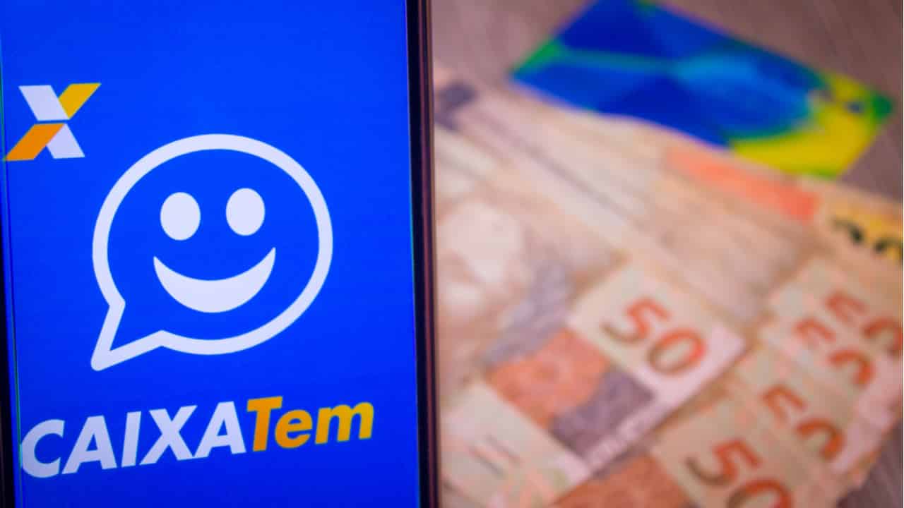 celular com tela aberta no aplicativo caixa tem, com notas de 50 reais e cartões de benefício atrás, represnetando o auxílio taxista.