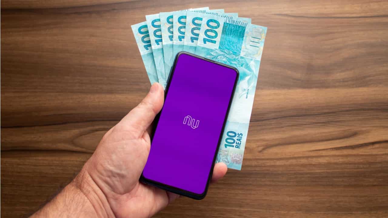 Celular com logo do Nubank em tela roxa. Ao fundo notas de 100 reais sobre uma superfície marrom
