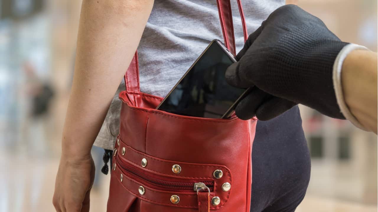 Mão vestida com luva preta furtando um aparelho celular de dentro de uma bolsa vermelha, carregada por uma pessoa no ombro.
