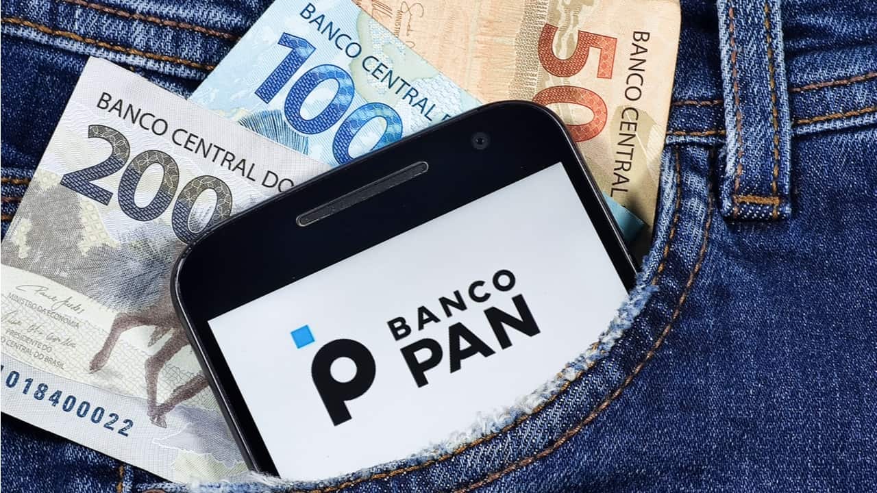 Imagem de um celular dentro de um bolso de calça jeans azul com o logotipo do Banco Pan na tela. Dentro do bolso, há também uma cédula de 200 reais, uma de 100 reais e outra de 50 reais
