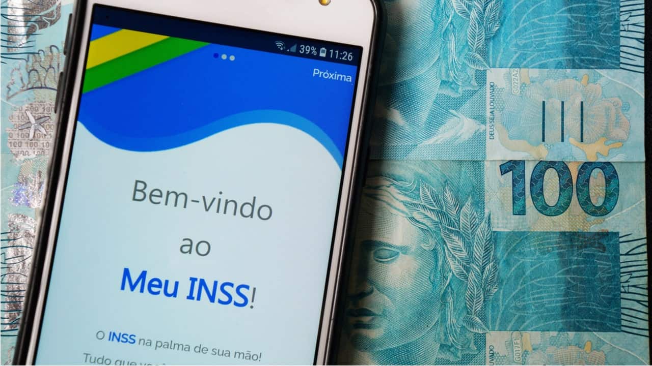 Celular com o aplicativo do INSS aberto, ao lado de uma nota de R$ 100,00