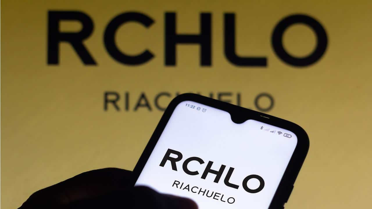 A o fundo logo da Riachuelo em marrom e preto, e em primeiro plano mão segurando celular com logo da marca Riachuelo em destaque na tela inicial