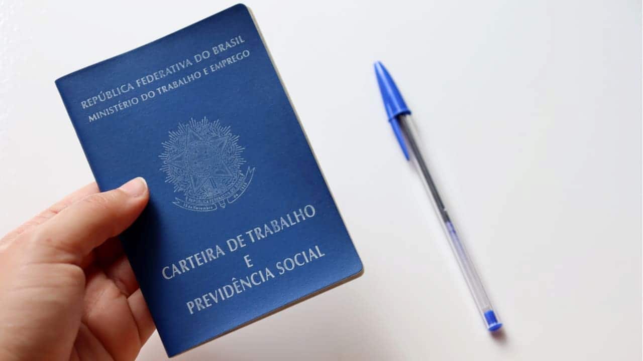 Uma mão segurando a carteira de trabalho e uma caneta azul sobre a mesa. leis trabalhistas