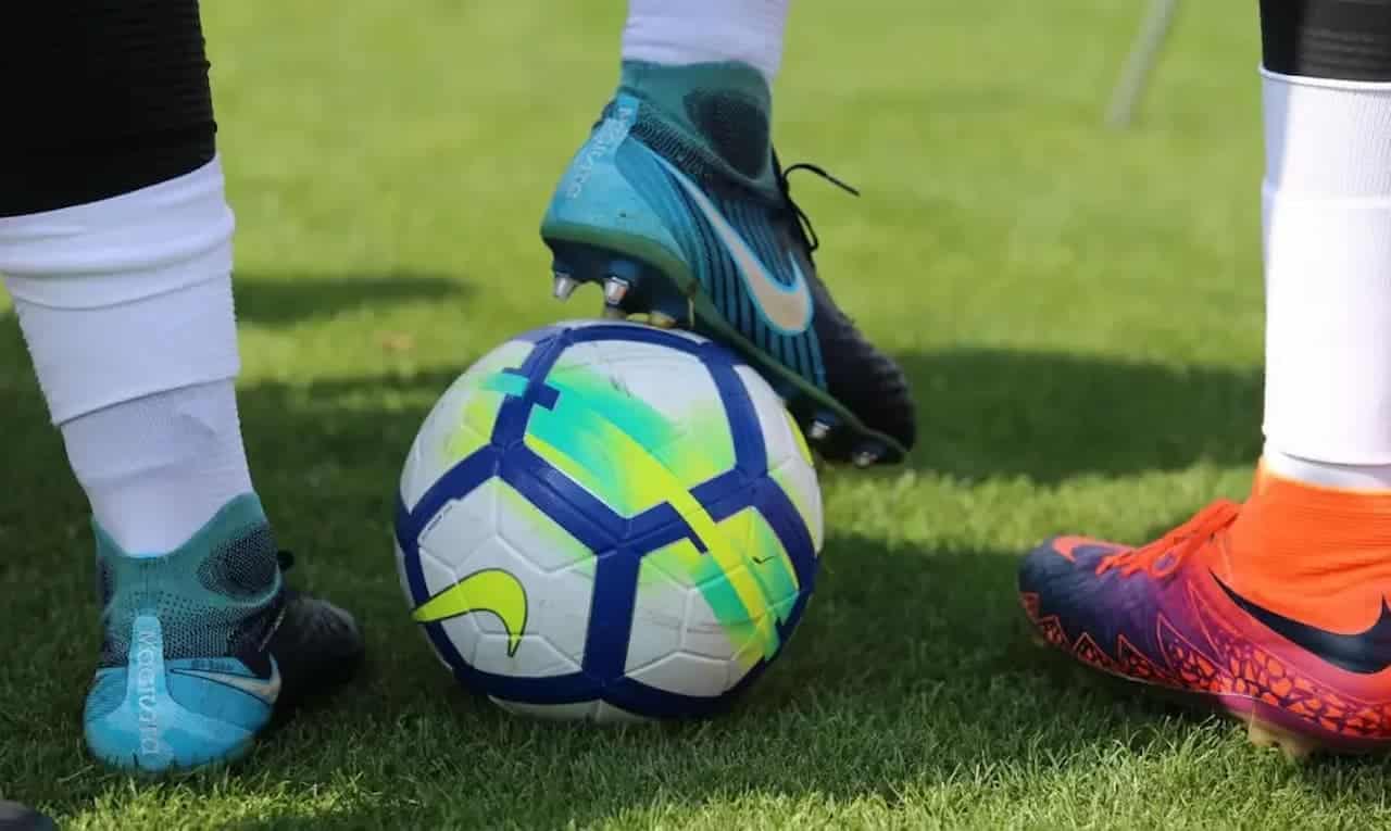 ingressos Na imagem aparecem os pés de um jogador de futebol parando uma bola. Ele usa chuteira da Nike preta e verde. No canto direito aparece o pé de outro jogador com chuteira laranja e rosa. cbf