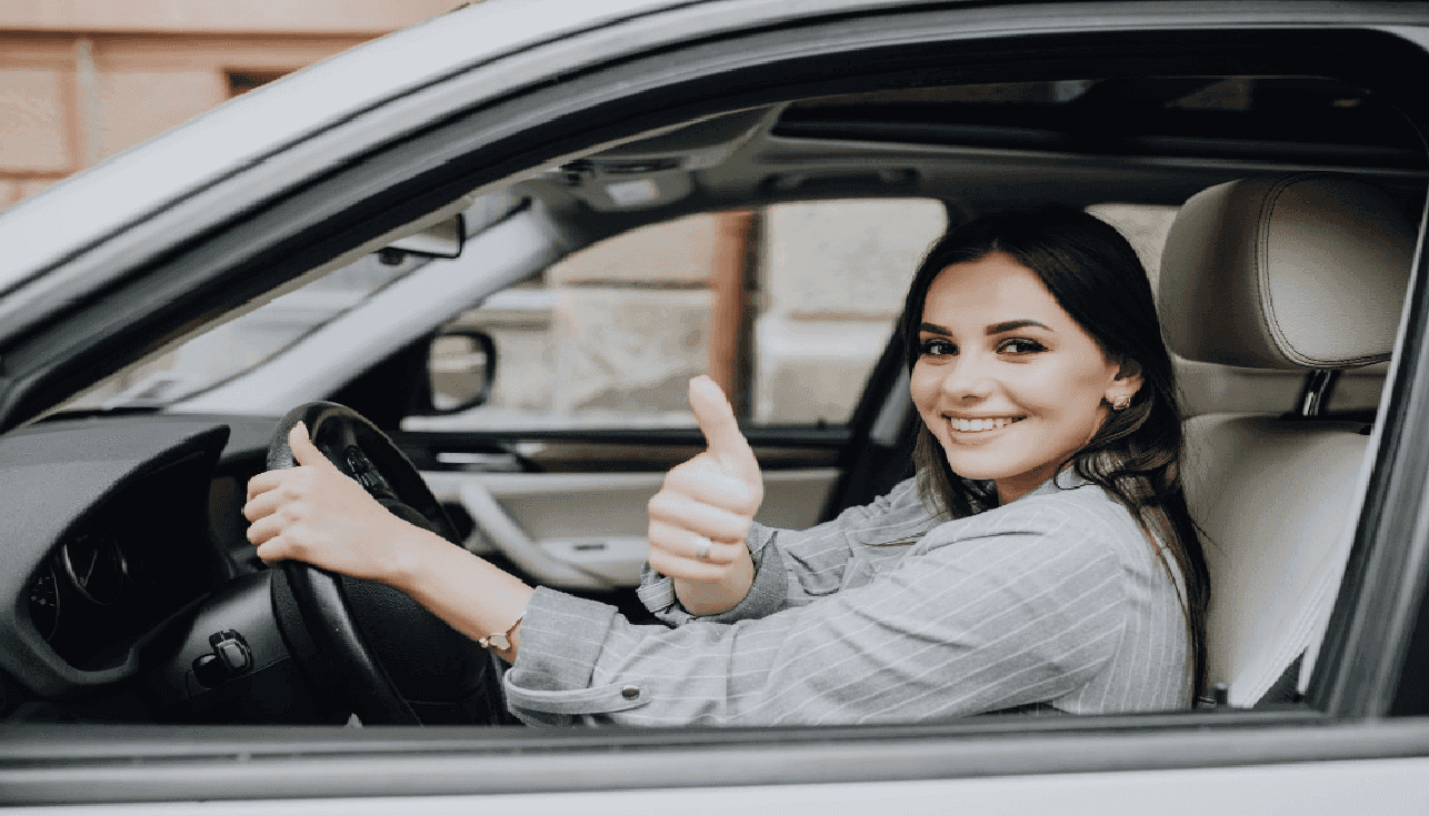 Moça em seu carro apoiando sua mãe esquerda no volante enquanto olha para a câmera fazendo um sinal de "legal" com a mão direita.