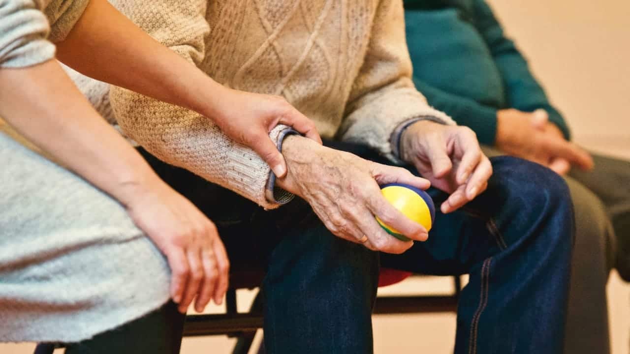 Imagem em que aparecem as mãos e o tronco de um idoso e de uma pessoa jovem; o idoso está segurando uma bolinha, a pessoa ao lado segura em seu braço