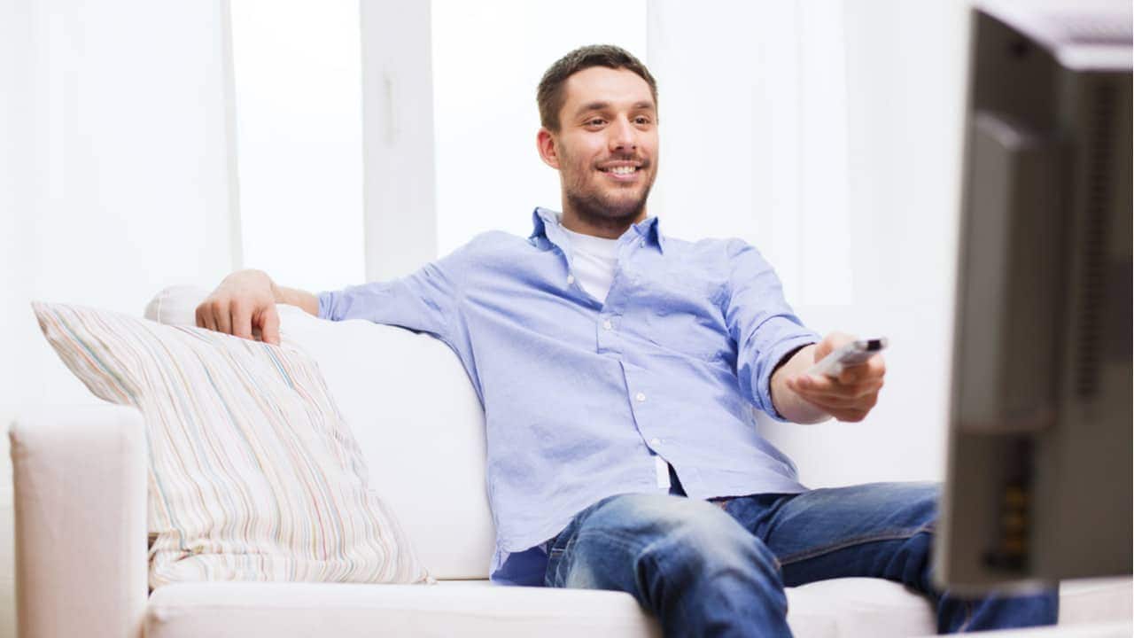 Homem sorridente sentado em sofá e apontando controle remoto para a televisão