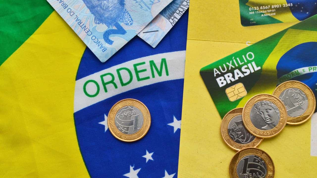 Cartão do Auxílio Brasil rodeado por notas de 2 reais e moedas de 1 real sobre uma bandeira do Brasil