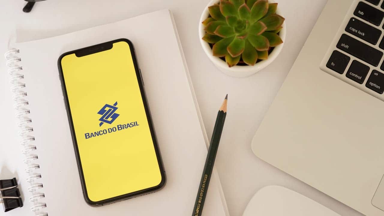 Aplicativo do Banco do Brasil na tela de um celular em cima de uma mesa, com um lápis, um caderno e uma planta pequena ao lado