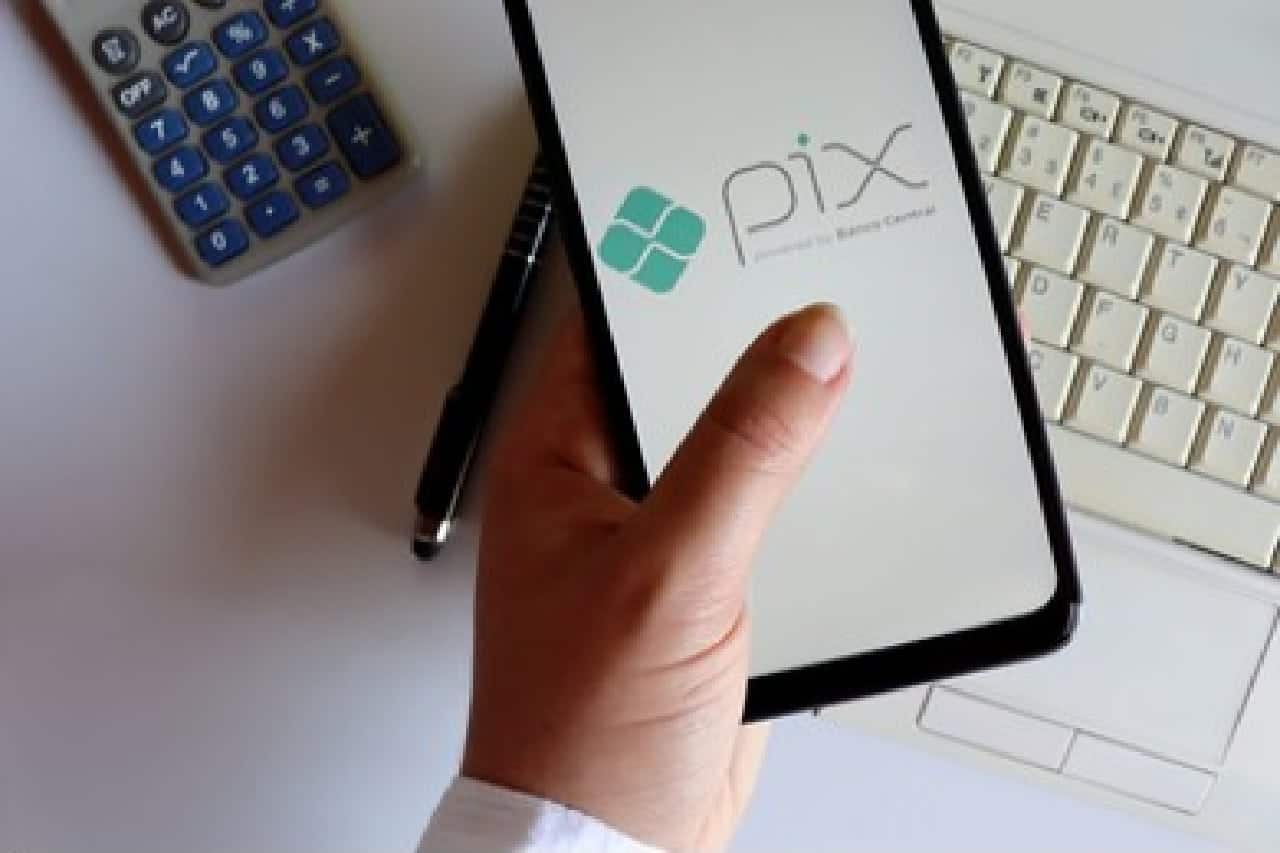 Mão segura smartphone com logo do Pix estampado na tela em cima de uma mesa em que há um teclado e uma calculadora também