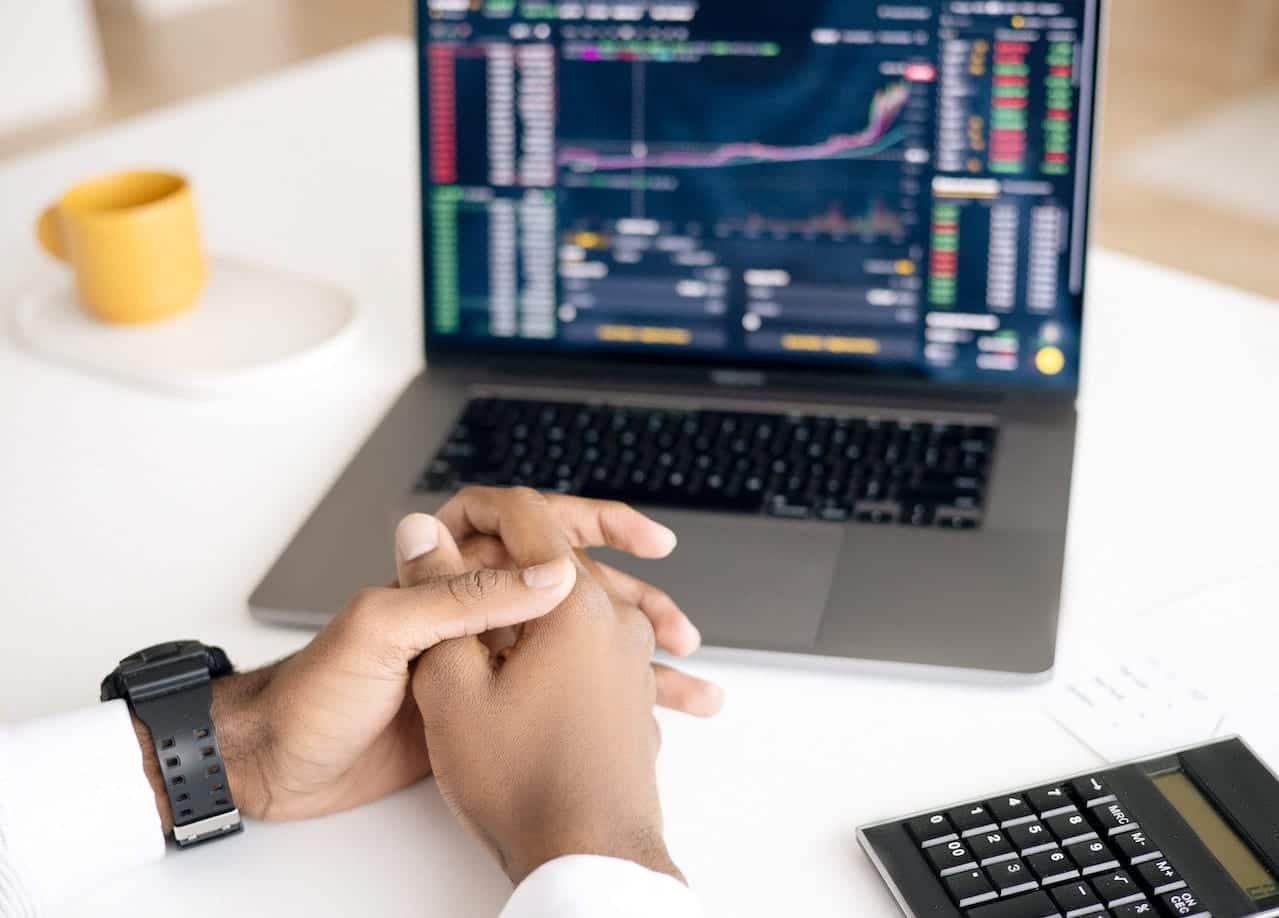 Mãos cruzadas em frente a um notebook que mostra índices do mercado financeiro em alusão a dividendos e JCP. Sobre a mesma mesa estão uma calculadora e uma xícara de café.