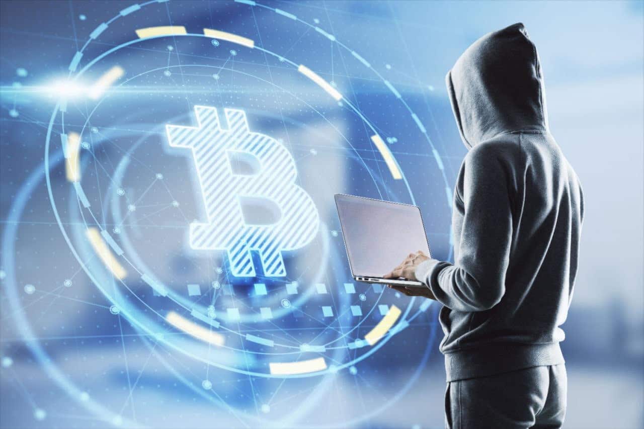 Pessoa vestida de preto, de costas, com capuz e notebook na mão, em frente a uma imagem digital com símbolo do Bitcoin