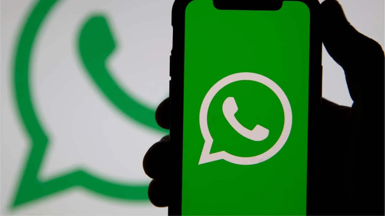 Mão segurando celular que mostra logo do WhatsApp. Ao fundo, outro logo do aplicativo aparece desfocado.