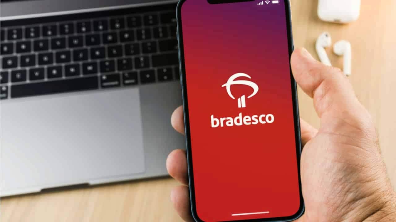Uma mão masculina com um celular utilizando o aplicativo do Bradesco, no fundo aparece um notebook