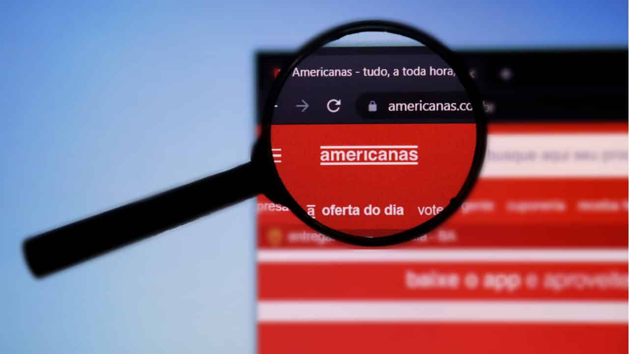 Lupa de aumento sobre a logo das Americanas enquanto o site da loja está aberto em uma tela de computador