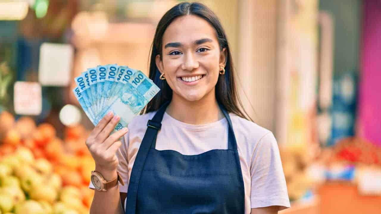 Mulher de avental, que aparenta ser empreendedora do ramo alimentício, está segurando notas de R$ 100 em formato de leque