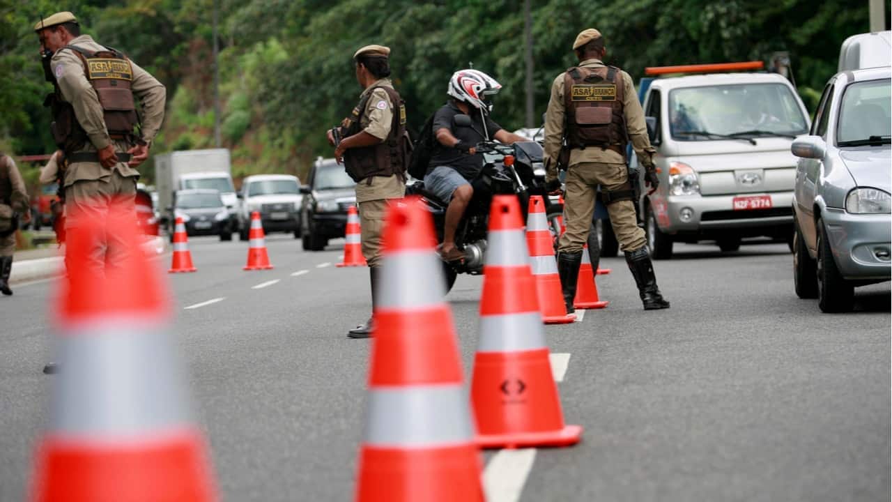Policias parados no meio de uma pista próximo de cones de sinalização, enquanto veículos se aproximam