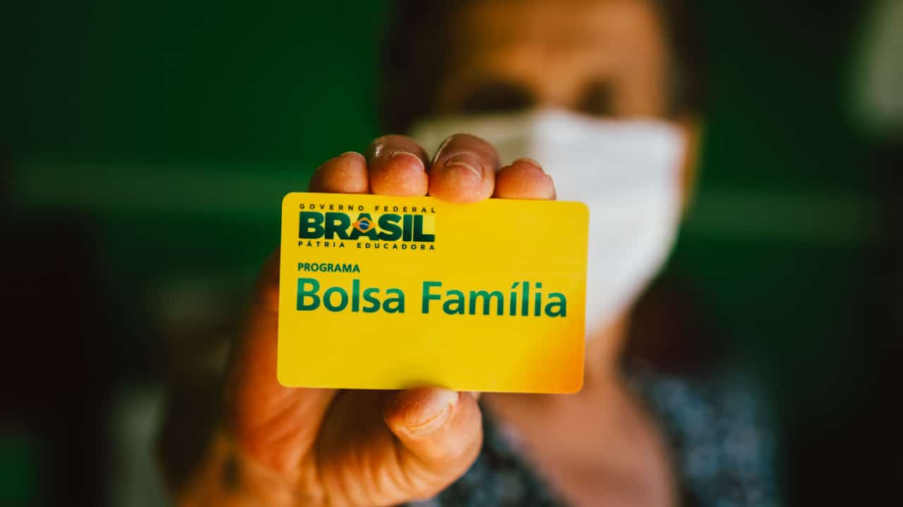 Mulher de máscara com rosto desfocado segurando cartão do programa Bolsa Família, que aparece em primeiro plano