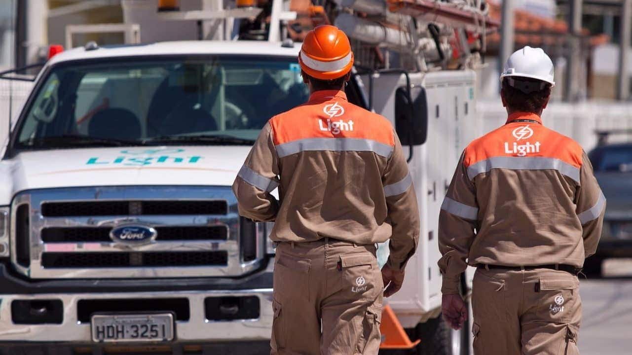 Dois funcionários uniformizados da empresa Light de costas indo em direção a um veículo com o logo da empresa