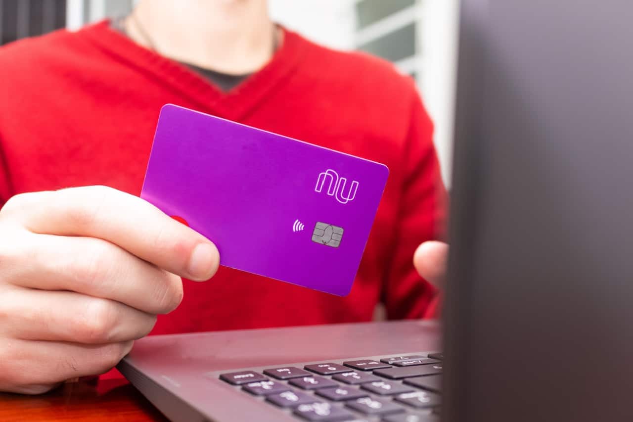 Homem de blusa vermelha usando um notebook enquanto segura cartão do Nubank