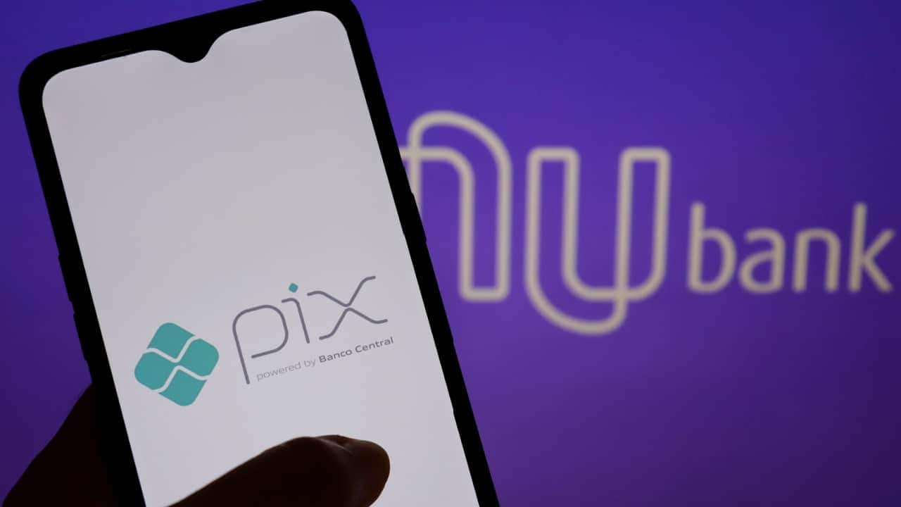 Mão segurando celular que mostra logo do Pix. Ao fundo, logo do Nubank aparece levemente desfocado.