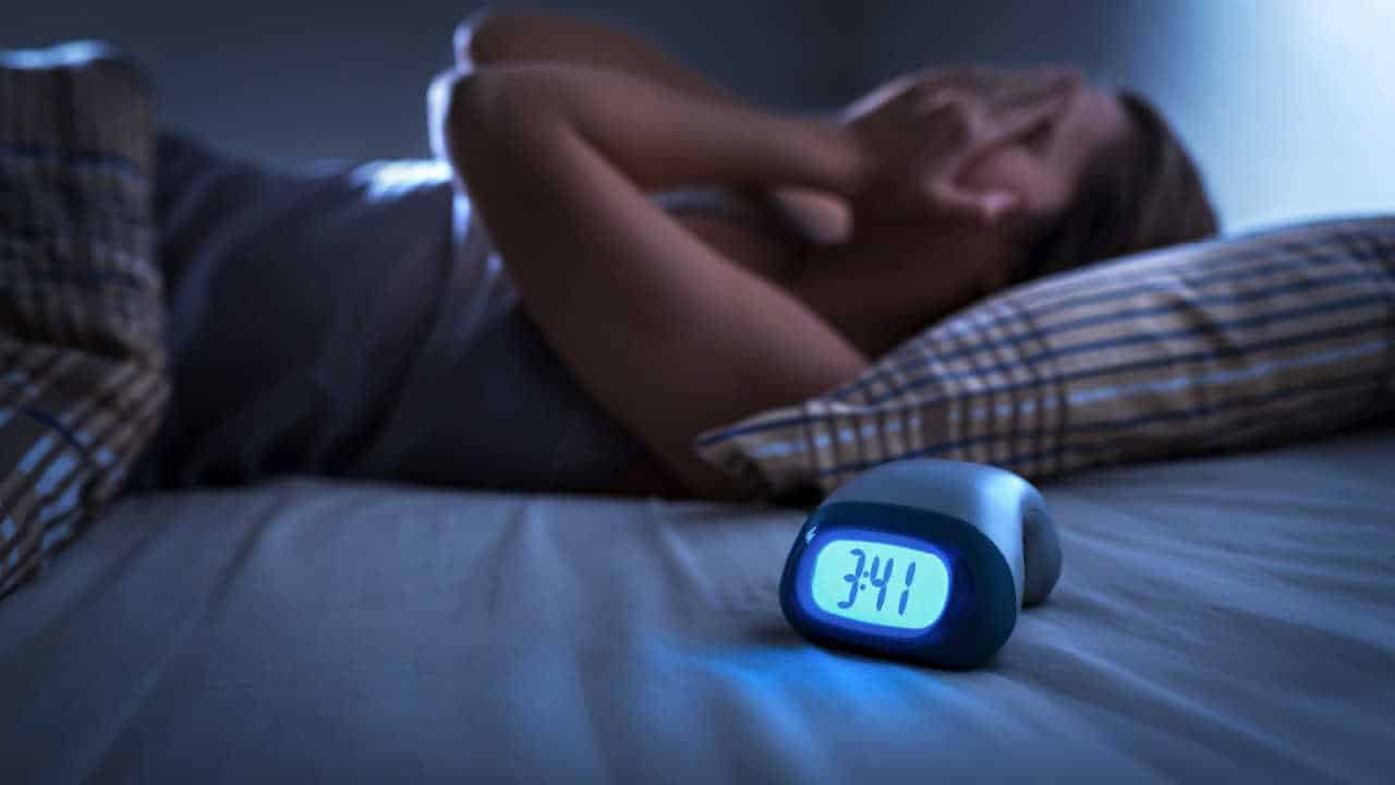 mulher deitada com as mãos no rosto pois não consegue dormir, ao lado de um relógio